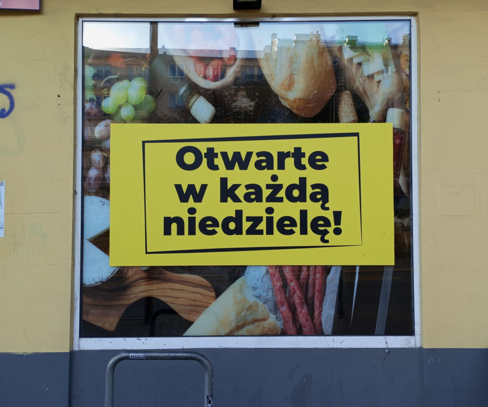 Żółta tablica z napisem Otwarte w każdą niedzielę! na tle witryny sklepowej, w której widoczne są grafiki produktów spożywczych, takich jak pieczywo, wędliny i owoce. Informuje o zasadach handlu w niedziele i możliwości robienia zakupów, o czym więcej przeczytasz na Super Biznes.