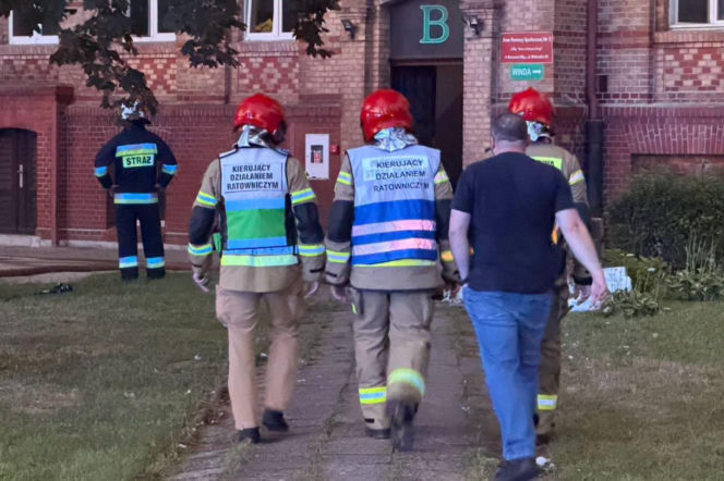 Strażacy w czerwonych hełmach i kamizelkach z napisem „Kierujący Działaniem Ratowniczym” na tle ceglanego budynku DPS w Gorzowie, gdzie miał miejsce pożar. Scena z akcji gaśniczej, o której więcej przeczytasz na naszym portalu.