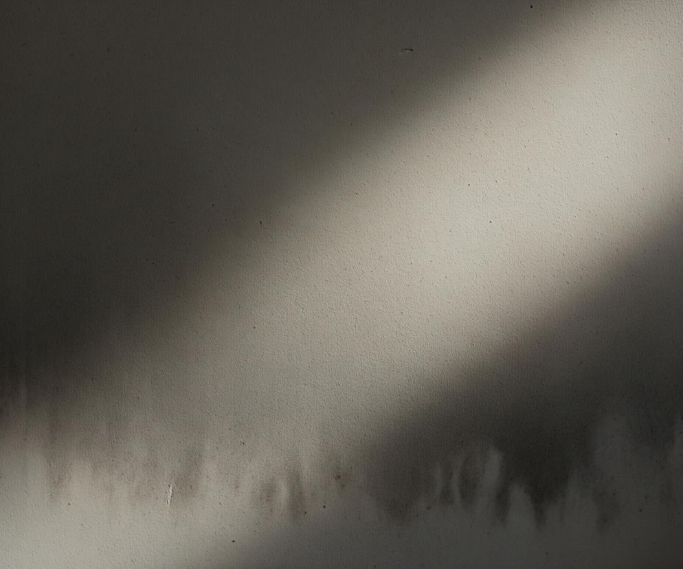 Lewa strona obrazu to zaciemniona, szara powierzchnia, stopniowo jaśniejąca w kierunku prawego dolnego rogu. Prawy górny róg jest rozjaśniony pasmem światła, biegnącym ukośnie w dół, tworząc jaśniejszy obszar na kremowej, teksturowanej ścianie. Na dole obrazu widoczne są nieregularne, ciemne zacieki, które rozprzestrzeniają się poziomo, przypominając kształtem niższe partie chmur. Całość ma ziarnistą fakturę i jest poprzecinana drobnymi, ciemnymi plamkami oraz pojedynczymi, białymi zadrapaniami.
