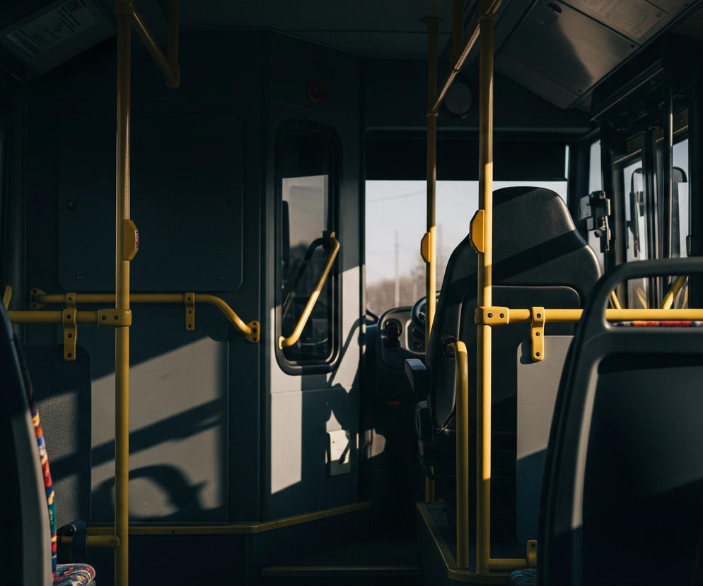 Wnętrze autobusu widoczne jest z perspektywy pasażera, zwróconego w kierunku przodu pojazdu. Przez duże okna widać jasne, bezchmurne niebo. Wnętrze charakteryzuje się szarymi ścianami i siedzeniami, z licznymi jasnożółtymi pionowymi i poziomymi poręczami. Światło słoneczne tworzy wyraźne cienie na ścianach i podłodze, a fotel kierowcy znajduje się po prawej stronie kadru, zasłaniając widok na przednią szybę.