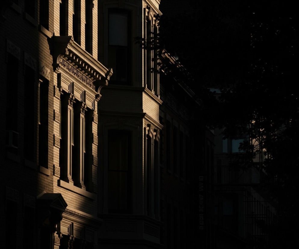 Lewa strona zdjęcia ukazuje fasadę ceglanego budynku z rzędem okien, częściowo oświetloną ciepłym, złotym światłem słonecznym. W centrum widoczny jest wykusz, którego lewa część jest również skąpana w świetle, podkreślając bogate detale architektoniczne, w tym zdobione gzymsy i parapety. Prawa strona obrazu zanurzona jest w głębokim cieniu, zarysy liści drzew częściowo zasłaniają ciemne, odległe konstrukcje.