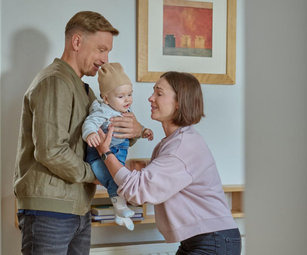 Igor (Jakub Wesołowski) trzyma niemowlę Jasia, ubranego w beżową czapkę i jeansy, patrząc na niego z uśmiechem, podczas gdy Anka (Sonia Mietielica) stoi obok, wyciągając ręce w stronę dziecka. Scena z serialu Na Wspólnej, o którym więcej przeczytasz na Super Seriale.