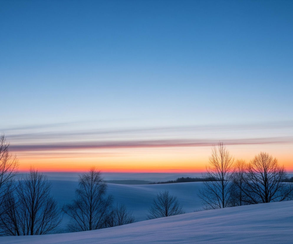 Zimowy krajobraz ukazuje pokryte śniegiem, pofalowane wzgórza, rozciągające się ku horyzontowi. Na pierwszym planie i nieco w głąb sceny widoczne są nagie drzewa liściaste o ciemnych, rozgałęzionych gałęziach, kontrastujące z bielą śniegu i jasnym niebem. Niebo, zajmujące około połowy kadru, przechodzi od ciemnoniebieskiego u góry, przez pasmo intensywnych barw pomarańczowo-czerwonych na horyzoncie, aż po jaśniejsze, błękitne odcienie tuż nad ośnieżonymi polami, z delikatnymi, poziomymi smugami chmur w środkowej części barwnej poświaty.