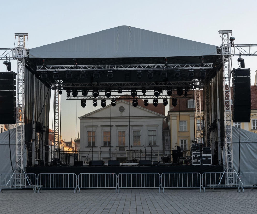 Na pierwszym planie widoczna jest duża, tymczasowa scena koncertowa, zadaszona szarym materiałem. Konstrukcja sceny składa się z metalowych rusztowań, na których zawieszone są liczne reflektory i systemy nagłośnieniowe w kolorze czarnym. Przed sceną rozstawiono szereg metalowych, srebrnych barierek ochronnych.

W tle, za sceną, widać budynki o różnobarwnych fasadach, z przewagą jasnych odcieni beżu i bieli, posiadające okna z ciemnymi framugami. Niebo jest bezchmurne, o jasnoniebieskiej barwie, z delikatnym, ciepłym blaskiem zachodzącego słońca widocznym za centralnym budynkiem. Podłoże jest wyłożone szarą kostką brukową.