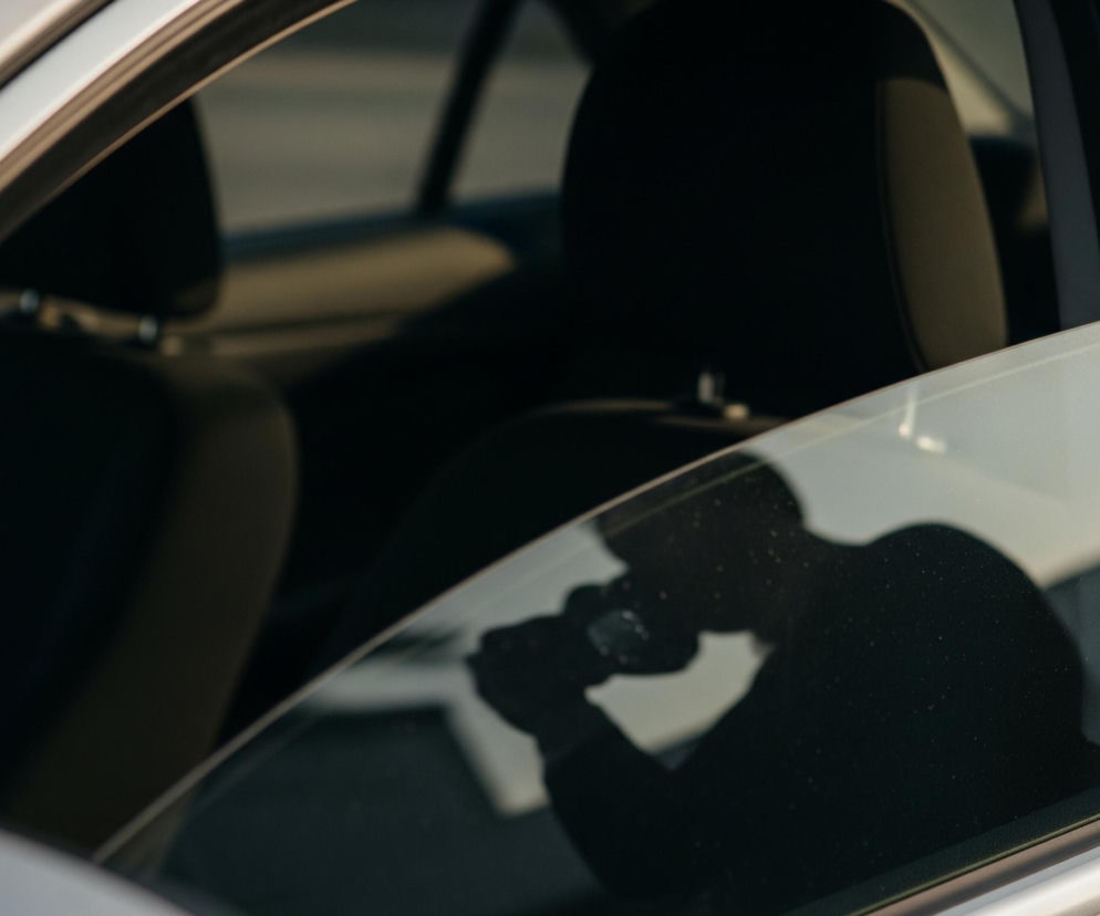 Częściowy widok na srebrny samochód, ukazujący lewą stronę karoserii i okna. W dolnej połowie zdjęcia, na szybie odbija się ciemna sylwetka osoby, która trzyma aparat fotograficzny, skierowany w stronę samochodu. W tle, za szybą, widać rozmazane, ciemne wnętrze pojazdu, w tym zagłówek fotela. Odbicie słońca pada na szybę, tworząc jasne refleksy, zwłaszcza w górnej części.