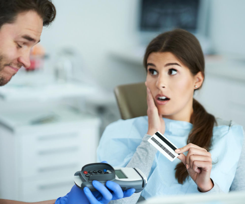 Młoda kobieta w fotelu dentystycznym trzyma kartę płatniczą z kodem kreskowym, patrząc z zaskoczeniem na terminal płatniczy podawany przez dentystę w rękawiczkach. Scena odzwierciedla obawy Polaków przed wysokimi rachunkami za leczenie stomatologiczne, o czym pisze Poradnik Zdrowie.
