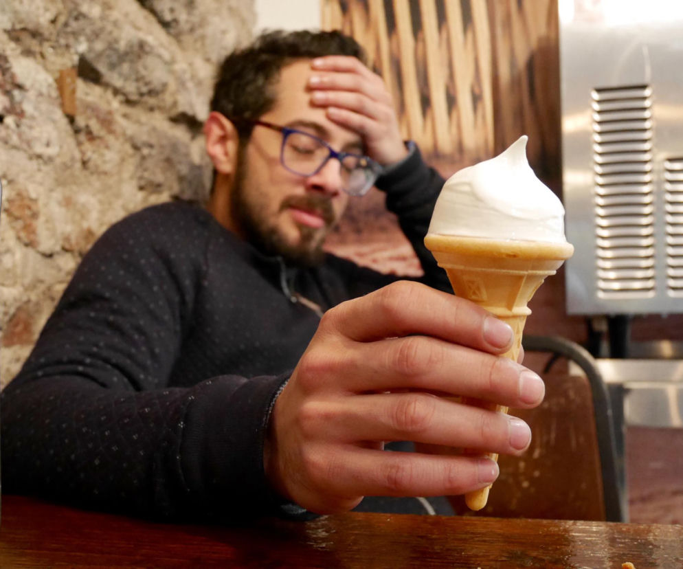 Mężczyzna z okularami, w czarnym swetrze, trzyma w ręku wafel z lodem, jednocześnie trzymając drugą rękę na czole w geście bólu głowy, tzw. zamrożenia mózgu. To ważny sygnał świadczący o skłonności do migren, o czym przeczytasz na Poradnik Zdrowie.