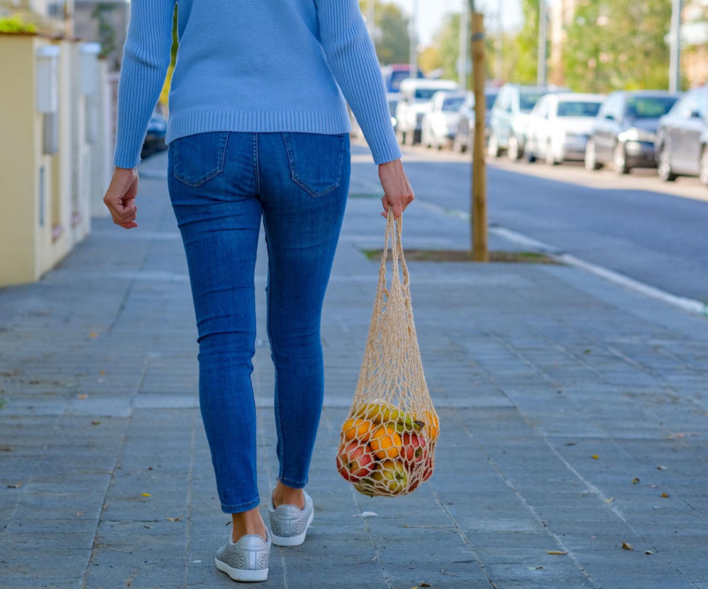 Kobieta niosąca siatkę z owocami w jednej ręce, idąca chodnikiem obok rzędu zaparkowanych samochodów. Ta codzienna czynność, niewinnie wyglądająca na zdjęciu, może prowadzić do poważnych problemów z kręgosłupem. Dowiedz się więcej o prawidłowym noszeniu zakupów na Poradnik Zdrowie.