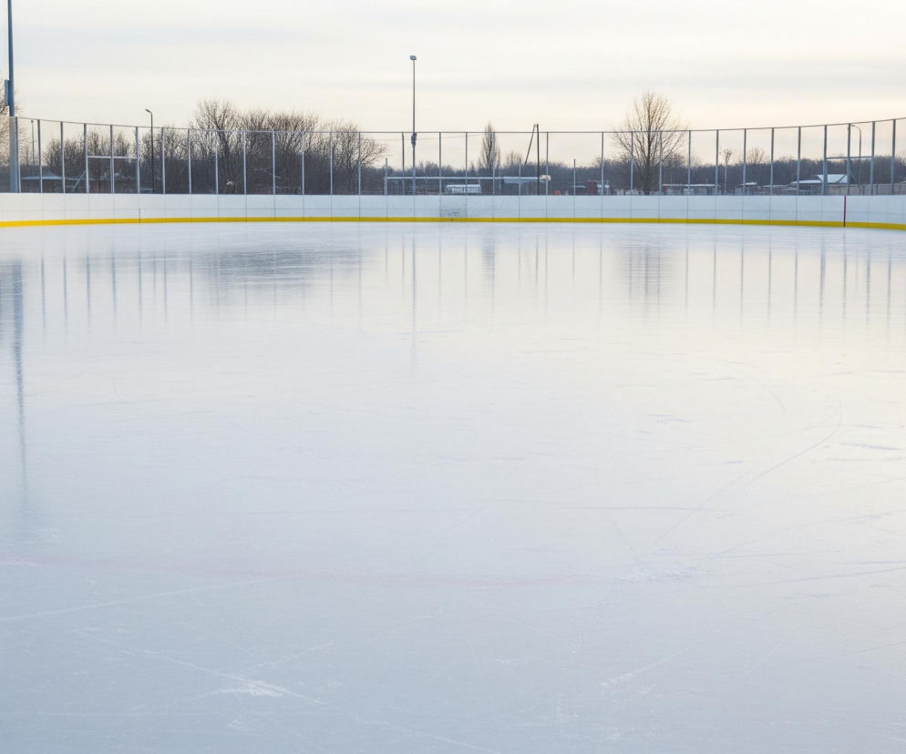 Duża, jasnoniebieska tafla lodu pokrywa większą część kadru, ze śladami łyżew widocznymi na jej powierzchni. Horyzontalna żółta linia biegnie wzdłuż górnej części lodu, oddzielając go od białego bandy. Pionowe czerwone i niebieskie linie zaznaczone są na bandzie, natomiast za bandą widać rząd siatek i nagich drzew. W tle widoczne są również słupy oświetleniowe i fragmenty budynków pod zachmurzonym, jasnym niebem.