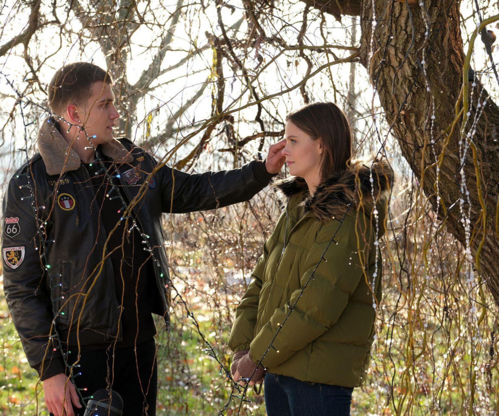 Młody mężczyzna w czarnej kurtce z futrzanym kołnierzem delikatnie dotyka policzka dziewczyny w zielonej kurtce, patrząc na nią z uczuciem. Scena z M jak miłość Mateusza i Majki, pełna romantyzmu i namiętności, dostępna na Super Seriale.