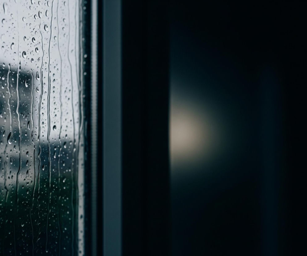 Na pierwszym planie widoczne jest szkło pokryte licznymi kroplami wody oraz ściekającymi smugami, co sugeruje deszcz. Za szkłem, w głębi, majaczy niewyraźny zarys szarego budynku i zieleń drzew. Po prawej stronie, obok pionowej ramy okiennej w kolorze ciemnoszarym, widoczne jest czarne, rozmyte tło z delikatną, owalną plamą światła w centralnej części.