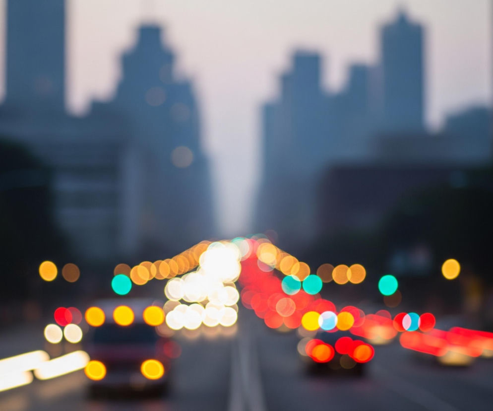 Centralny element obrazu to rozmyty, nocny lub wieczorny pejzaż miejski, z wyraźnie widocznymi światłami samochodów tworzącymi smugi świetlne. Na pierwszym planie, w dolnej części, znajduje się droga z biało-żółtymi smugami światła od przednich reflektorów samochodów jadących w kierunku widza po lewej stronie, oraz czerwonymi smugami od tylnych świateł pojazdów oddalających się po prawej stronie. Liczne, kolorowe, okrągłe plamy światła bokeh, w odcieniach żółci, pomarańczy, czerwieni i zieleni, rozciągają się wzdłuż drogi w kierunku horyzontu. Tło stanowią rozmazane sylwetki wysokich budynków w kolorze ciemnego błękitu i szarości, pod rozświetlonym, jasnoszarym niebem.