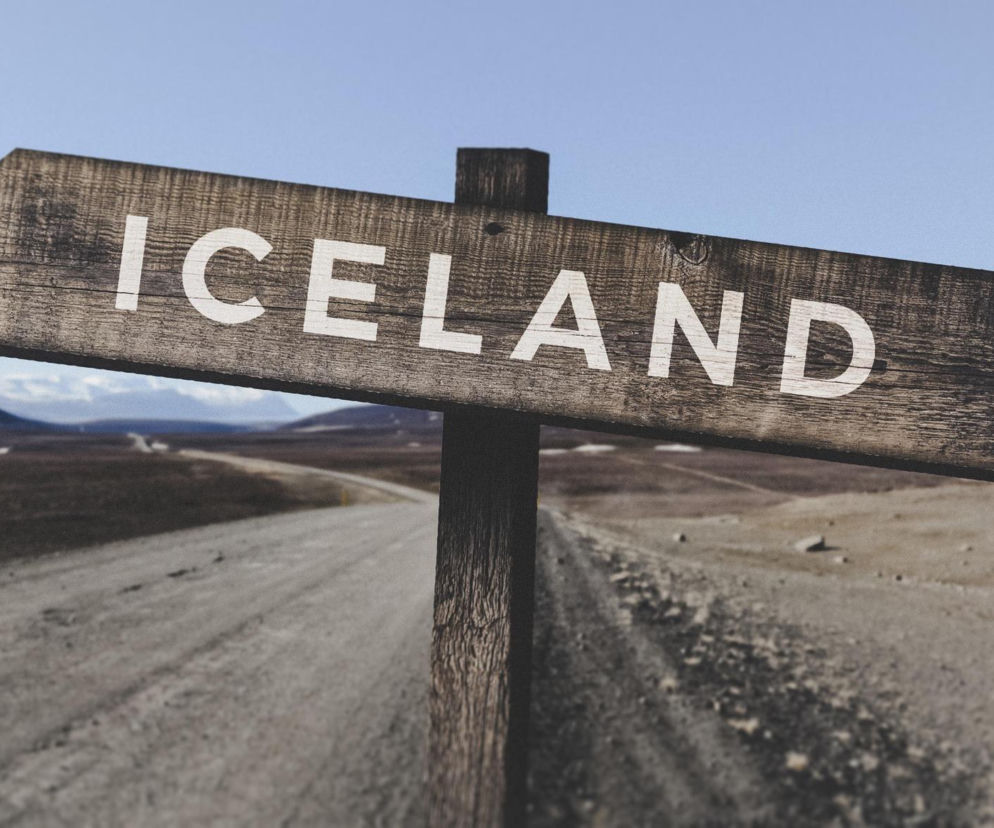 Drewniany drogowskaz z napisem ICELAND na tle surowego krajobrazu. O lotach z Gdańska na Islandię przeczytasz w Eskce.