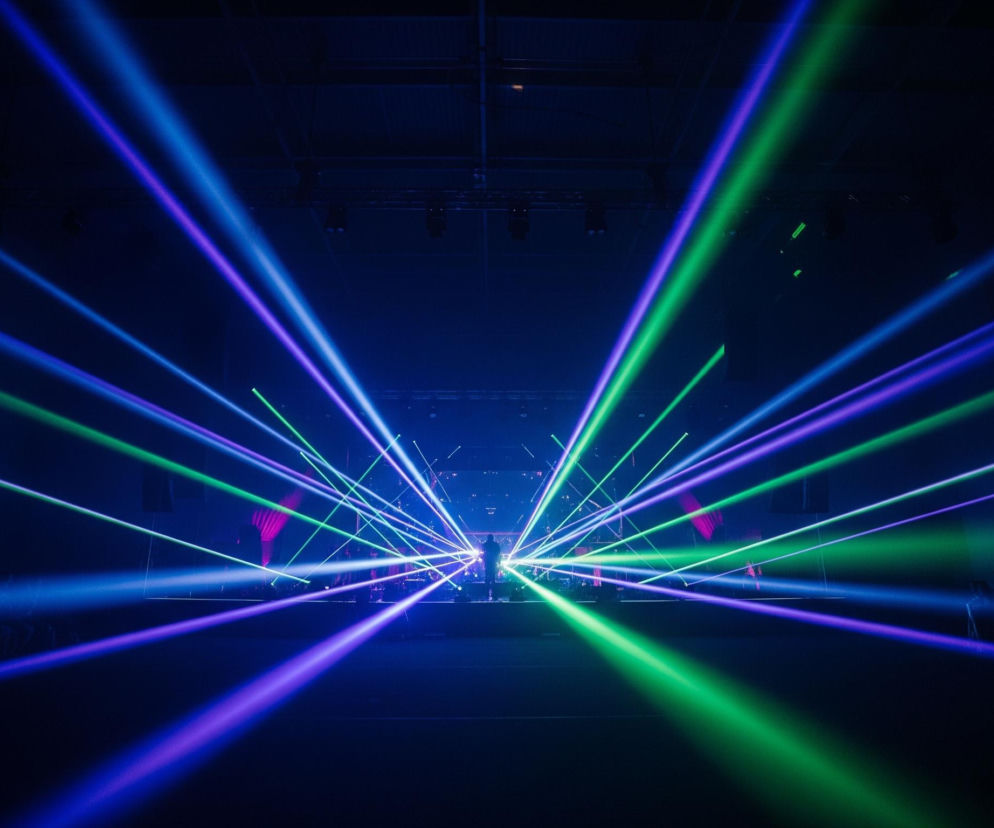 Głębokie ciemności wypełnia wielka sala, w której krzyżują się liczne, intensywnie niebieskie, zielone i fioletowe promienie światła. Światła wychodzą z centralnej sceny na horyzoncie, rozciągając się dynamicznie w kierunku widza i tworząc w powietrzu skomplikowaną sieć świetlnych smug. W oddali na scenie widoczne są niewyraźne sylwetki ludzi oraz sprzętu, a po bokach sceny zarysowują się podłużne głośniki. W dolnej części obrazu, pod promieniami światła, widać rozmyte kontury publiczności, która wydaje się unosić ręce w górę.