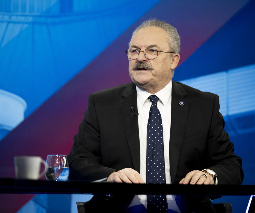 Portret polityka Marka Jakubiaka z wąsem i w okularach, ubranego w garnitur i krawat w kropki, siedzącego przy stole studyjnym. W tle widoczne są niebiesko-czerwone elementy scenografii, odzwierciedlające kontekst jego wypowiedzi o lekceważeniu Polski przez Zełenskiego, o których przeczytasz na naszym portalu.