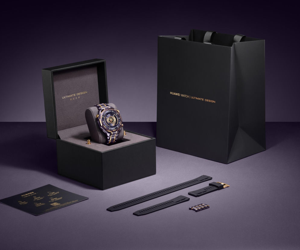 Luksusowy smartwatch Huawei Watch Ultimate Design Royal Gold Edition w otwartym pudełku, obok czarna torba prezentowa, instrukcja i dodatkowe paski. Zegarek ze złotymi akcentami symbolizuje bogactwo. Więcej o produkcie na Super Biznes.