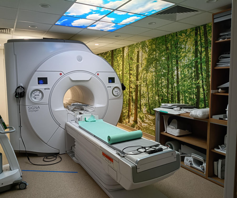 Biały rezonans magnetyczny GE SIGNA Voyager w nowoczesnym szpitalu urologicznym w Ząbkach, otoczony fototapetą z lasem i podświetlanym sufitem imitującym niebo. Sprzęt pozwala na precyzyjną diagnostykę raka, o czym możesz przeczytać na Poradnik Zdrowie.