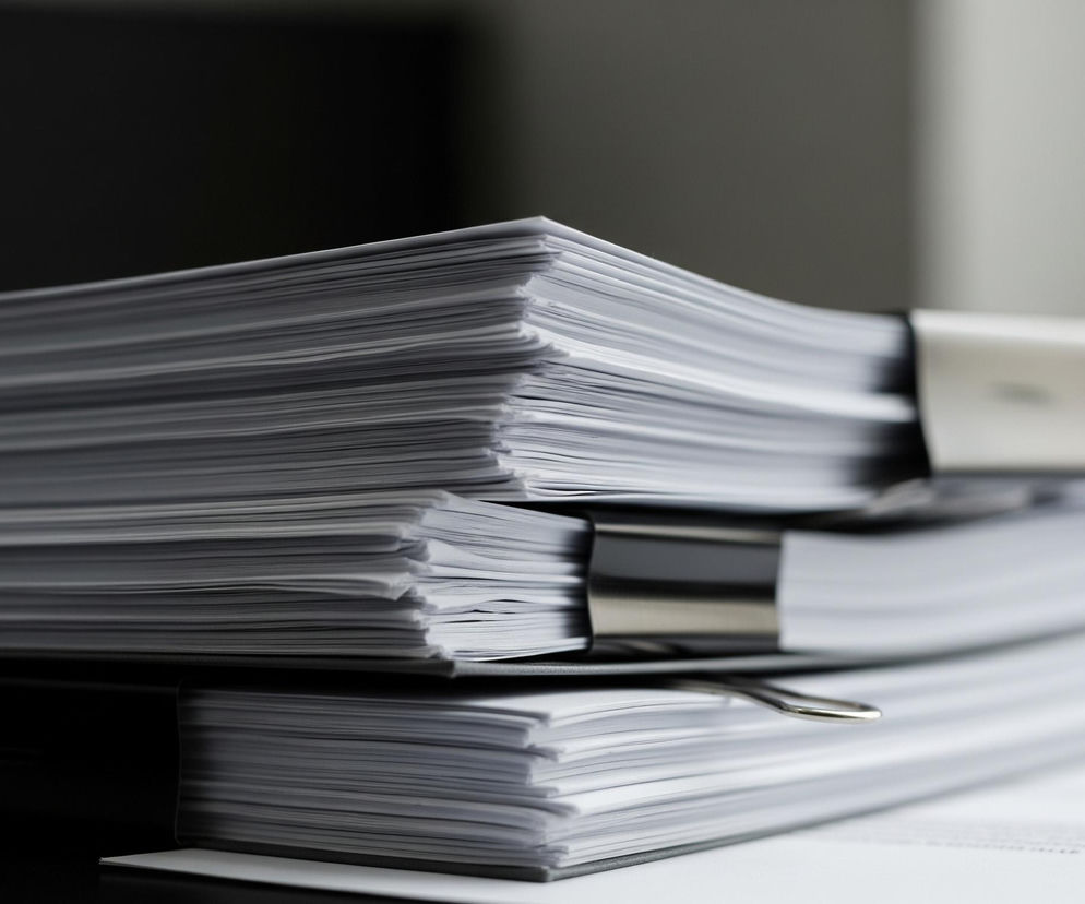 Na pierwszym planie widoczne są trzy stosy białych kartek papieru, ułożone jeden na drugim. Każdy stos jest zaciśnięty czarnym, metalowym klipsem biurowym, utrzymującym razem poszczególne kartki. Brzegi papieru są lekko nierówne, co nadaje im teksturę i wrażenie naturalnego zużycia. Tło jest rozmazane, przechodząc od ciemnoszarego po lewej stronie do jasnoszarego po prawej.