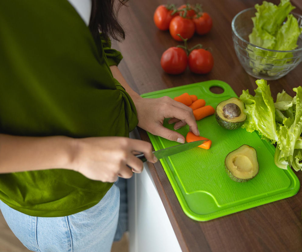 Kobieta w zielonej bluzce kroi marchew na jasnozielonej plastikowej desce do krojenia, obok leżą awokado i sałata. Artykuł na Poradnik Zdrowie wyjaśnia, dlaczego plastikowe deski mogą być siedliskiem bakterii i mikroplastiku.