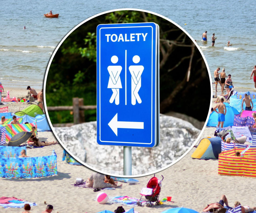 Niebieska tablica kierunkowa „TOALETY” z białymi piktogramami mężczyzny i kobiety, osadzona na piaszczystej plaży w Kołobrzegu, pełnej turystów, parawanów i namiotów, którzy opalają się lub kąpią w morzu. O karach dla turystów przeczytasz na Super Biznes.