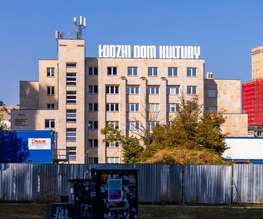 Gmach Łódzkiego Domu Kultury z jasną elewacją za metalowym płotem placu budowy. O zabytku przeczytasz na Architektura.