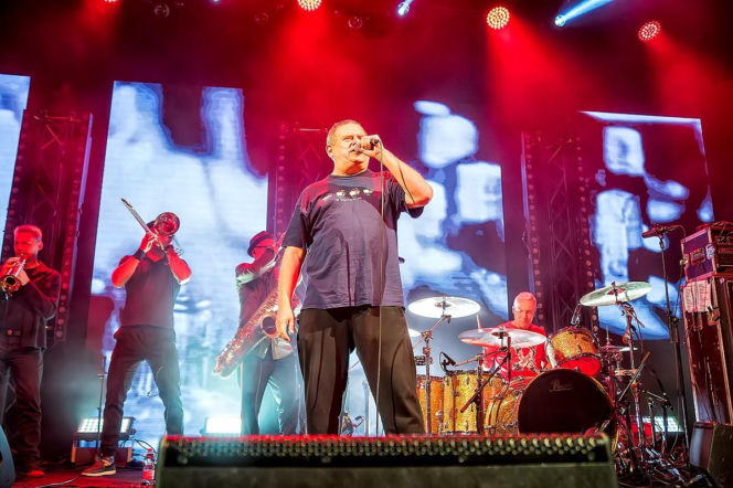 Koncert zespołu Kult, w centrum kadru Kazik Staszewski śpiewający do mikrofonu, ubrany w granatową koszulkę i ciemne spodnie. Za nim pozostali muzycy: trębacz, puzonista, saksofonista oraz perkusista, oświetleni jaskrawym, czerwonym i niebieskim światłem scenicznym. Więcej o problemach zdrowotnych lidera Kultu można przeczytać na portalu Poradnik Zdrowie.