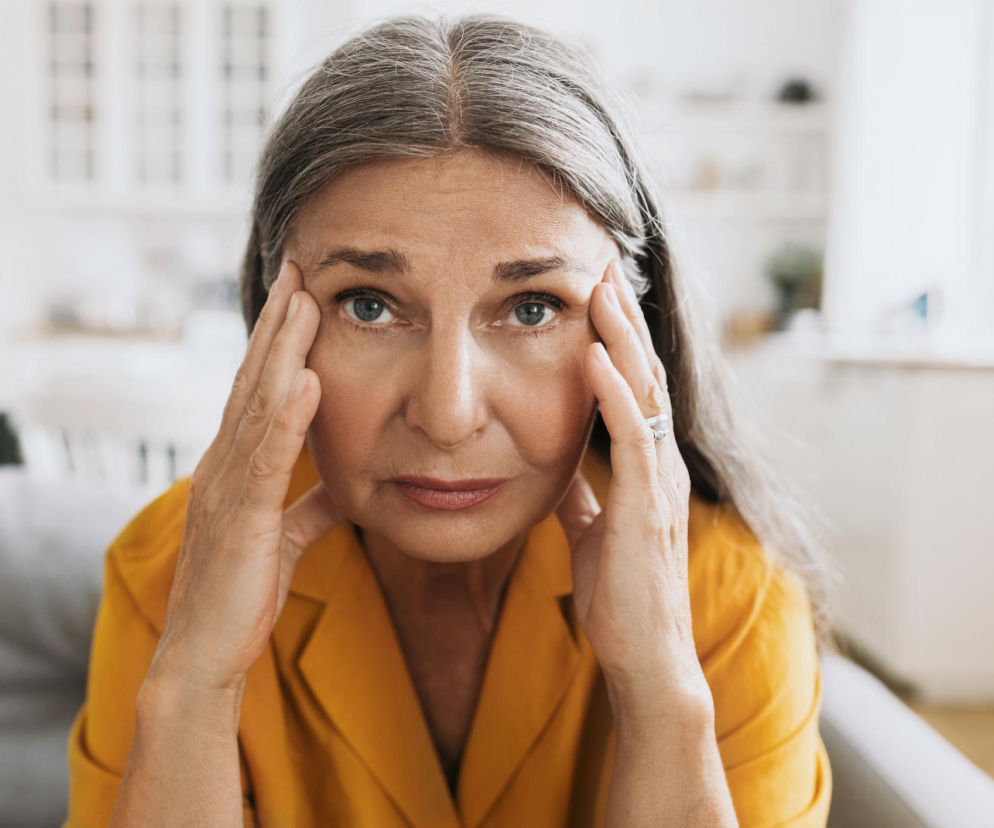 Starsza kobieta z siwymi włosami trzymająca dłonie przy skroniach, z zatroskanym wyrazem twarzy, nawiązująca do problemów z pamięcią i skupieniem podczas menopauzy, o których przeczytasz więcej na Poradnik Zdrowie.