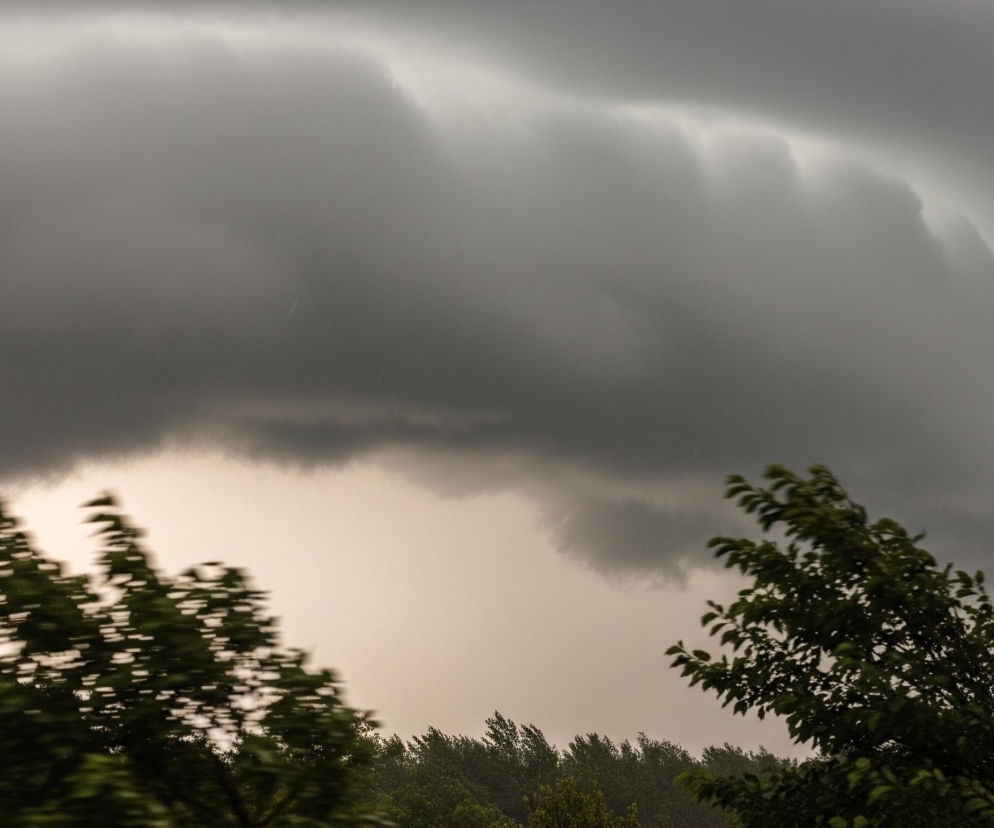 Na obrazie dominuje zachmurzone, ciemnoszare niebo zajmujące większą część kadru. W górnej części widoczne są warstwowe, pofalowane formacje chmur, z jaśniejszymi fragmentami wskazującymi na źródło światła lub inną strukturę. Pod chmurami, w centralnej części zdjęcia, niebo ma jaśniejszy, bladoszary odcień, a pojedyncze krople deszczu są dostrzegalne. Na pierwszym planie i po bokach widać rozmyte, zielone korony drzew, sugerujące silny wiatr lub ruch.