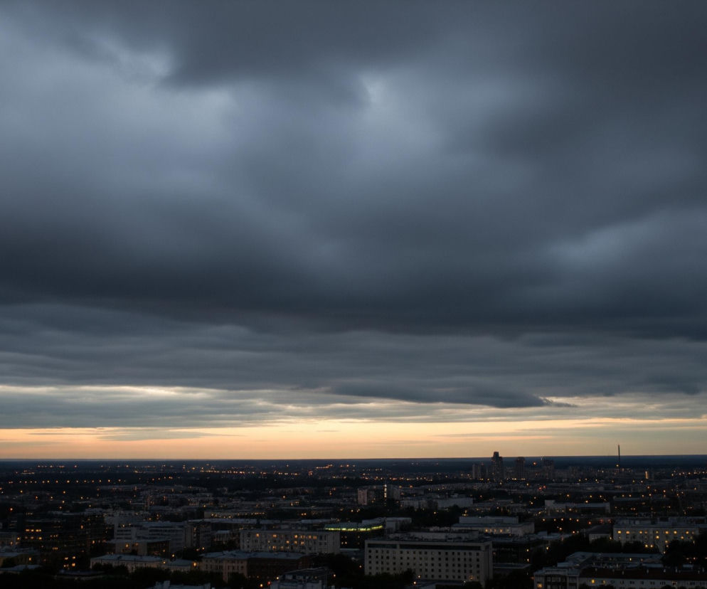 Niebo zajmujące około dwie trzecie górnej części obrazu jest pokryte gęstymi, ciemnoszarymi chmurami, z których niektóre mają jaśniejsze, szare akcenty, szczególnie w dolnej części, gdzie przechodzą w delikatne pasmo jasnego, lekko pomarańczowego światła. Poniżej rozciąga się miejski krajobraz, który w dużej mierze jest pogrążony w ciemności, z licznymi punktami jasnego, żółto-pomarańczowego światła pochodzącymi z okien budynków i latarni ulicznych. Wśród zabudowy widoczne są bloki mieszkalne i wieżowce, a na horyzoncie zarysowują się sylwetki dalszych budynków i pojedynczy komin, wszystko to pod ciemnym, pochmurnym niebem o zmierzchu.