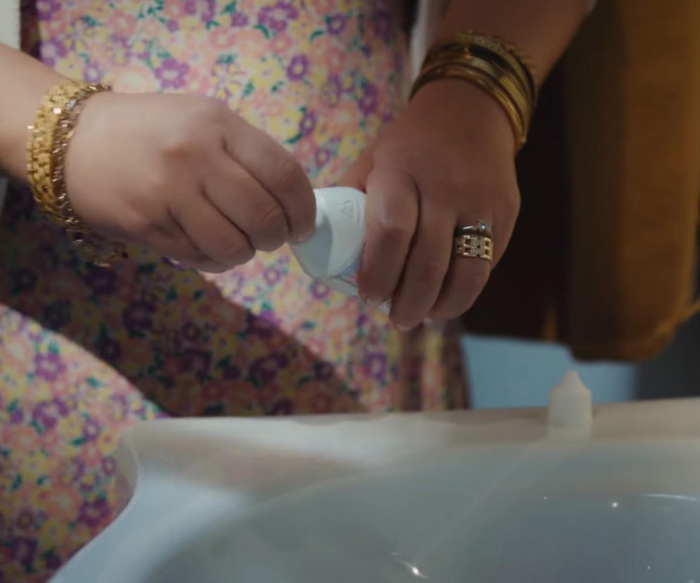 Bliskie ujęcie rąk kobiety w kolorowej kwiecistej sukience, z licznymi złotymi bransoletami i pierścionkami, przelewającej płyn z białej buteleczki nad umywalką. Scena przedstawia podstępny czyn Cansel z serialu Dziedzictwo, o którym przeczytasz na Super Seriale.