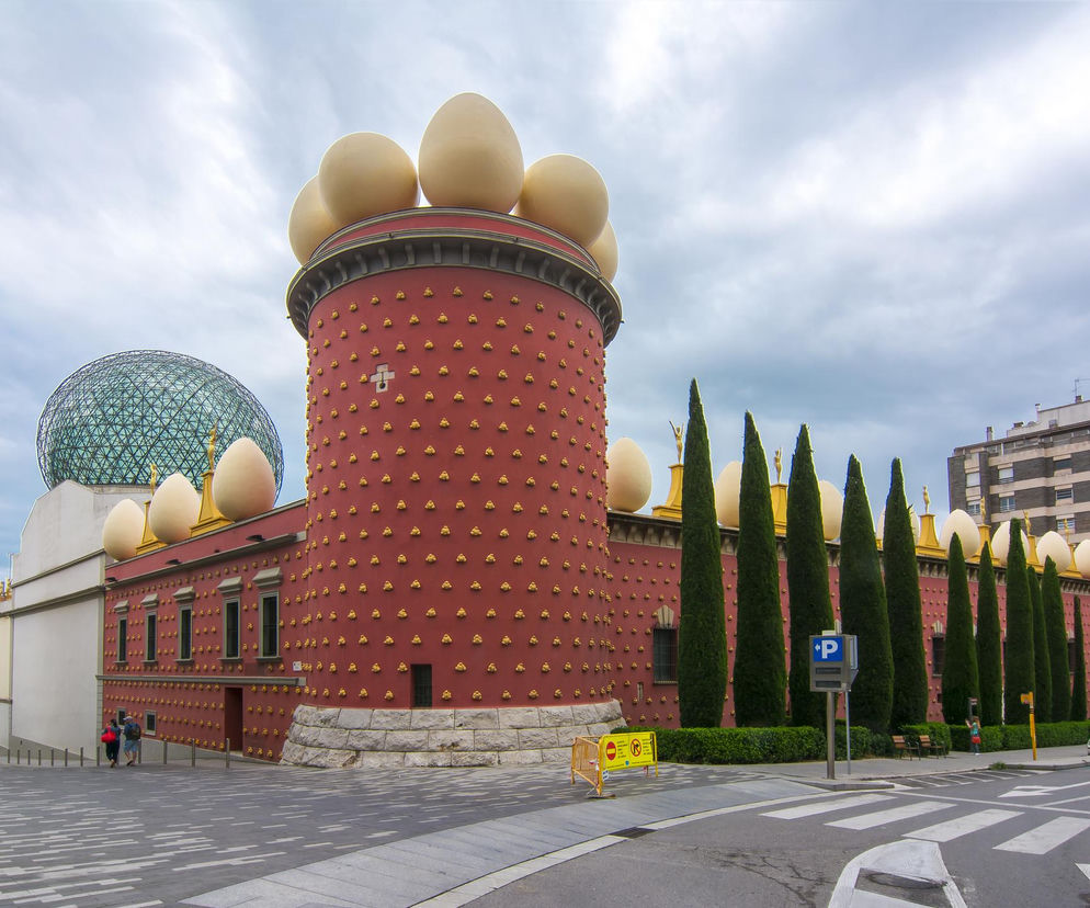 Muzeum Salvadora Dalego w Figueres, Hiszpanii, z fasadą w kolorze terakoty ozdobioną gipsowymi bochenkami chleba i ogromnymi jajami na dachu. W tle widoczna przeszklona kopuła, a obok strzeliste cyprysy. O surrealistycznej architekturze tego niezwykłego miejsca przeczytasz na Architektura Murator Plus.