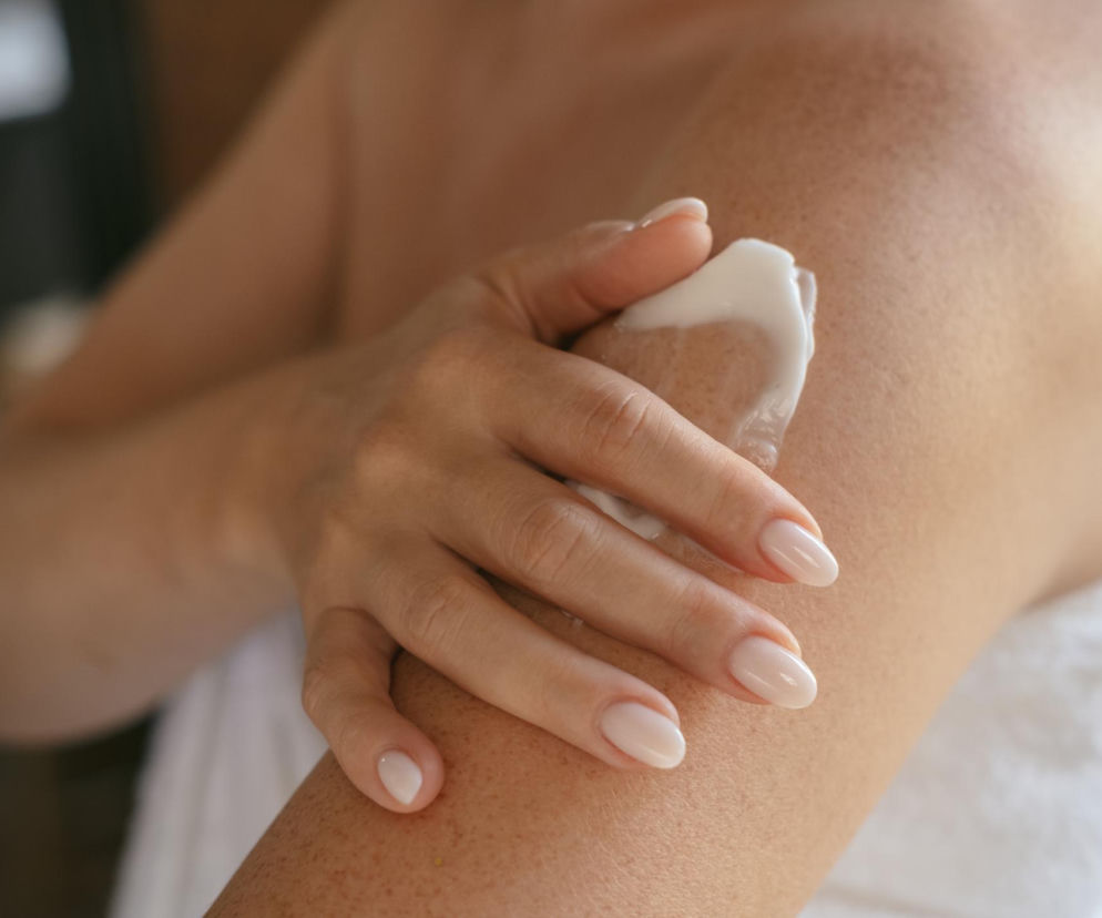 Dłoń kobiety w delikatnym, jasnym manikiurze rozsmarowująca biały krem na swoim ramieniu, symbolizująca pielęgnację podrażnionej skóry. Zdjęcie ilustruje znaczenie odpowiedniej pielęgnacji ciała, szczególnie po radioterapii, o czym przeczytasz na Poradnik Zdrowie.