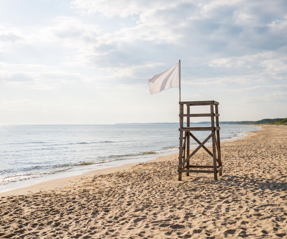 Drewniana wieża ratownika z białą flagą na pustej plaży w Mrzeżynie. O warunkach nad morzem dowiesz się z SE Szczecin.