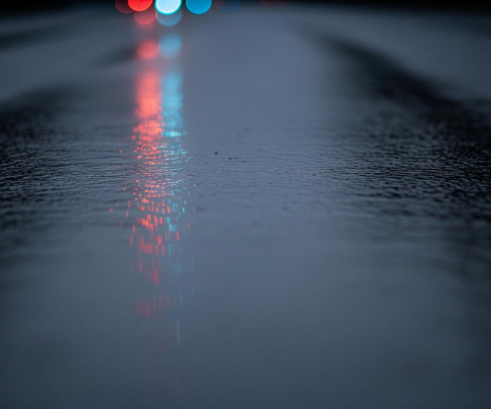 Ciemna, mokra nawierzchnia drogi z widocznymi odbiciami świateł w górnej części kadru. Odbicia tworzą pionowe smugi, głównie w odcieniach czerwieni i turkusu, które rozmywają się w kierunku dołu. Powierzchnia drogi ma nierówną teksturę, co sugeruje kałuże lub deszczową pogodę. Cały obraz jest utrzymany w ciemnych, chłodnych barwach, z dominującymi odcieniami szarości i granatu, a światła dodają mu kontrastu.