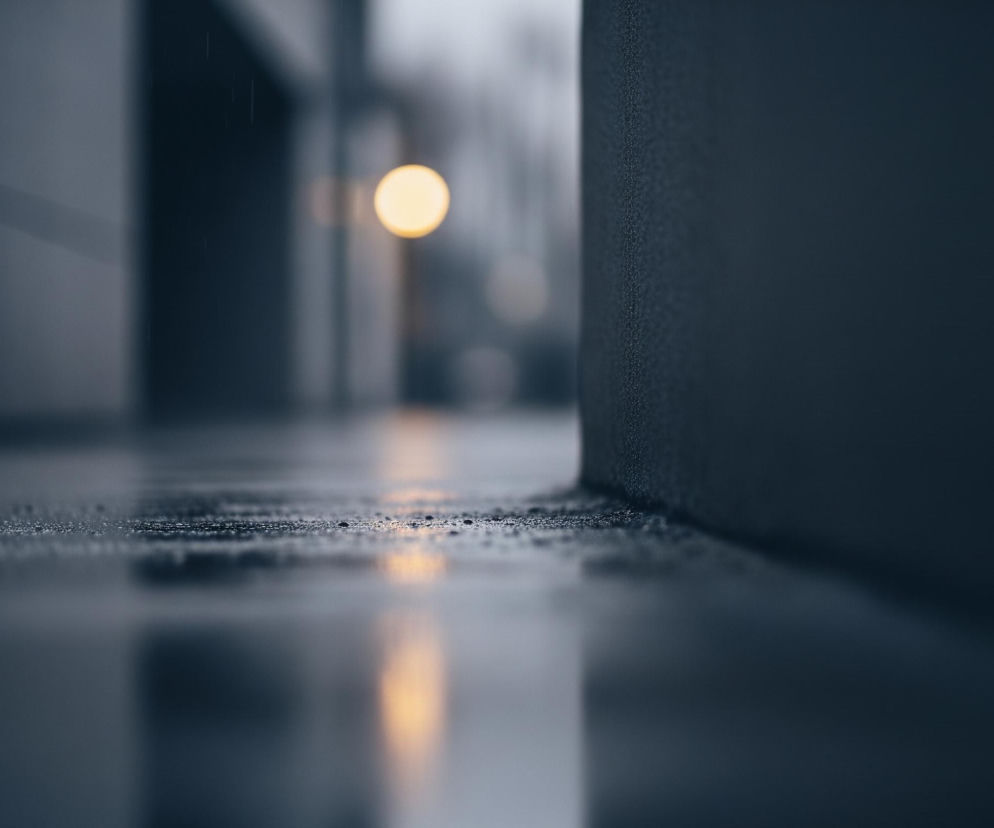 Szyba lub mokra powierzchnia znajduje się na pierwszym planie, pokryta drobnymi kroplami deszczu. Po prawej stronie wznosi się ciemna, teksturowana ściana, natomiast po lewej stronie widać odbicie innej, jasnej ściany z poziomym pasem. W tle, rozmyte żółte światło, przypominające lampę uliczną, odbija się w kałuży na pierwszym planie, tworząc pionowy, rozciągnięty blask. Całość utrzymana jest w chłodnych, ciemnych odcieniach szarości i błękitu.