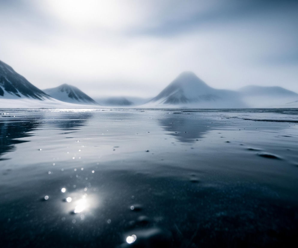 Na pierwszym planie rozciąga się ciemna, płytka woda lub mokry lód, odbijająca jasne punkty światła, tworzące efekt bokeh. Dalej, za linią brzegową, wznoszą się ośnieżone, spiczaste góry, częściowo zasłonięte przez zamglone, jasne niebo. Cała scena utrzymana jest w chłodnych odcieniach błękitu, szarości i bieli, co nadaje jej surowy, zimowy charakter.