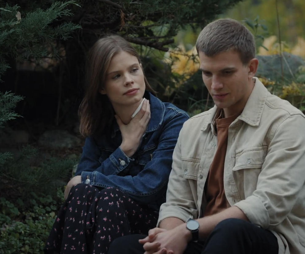 Młodzi Mateusz i Majka, siedzą w plenerze wśród zieleni, rozmawiając o życiu uczuciowym. On w beżowej koszuli, ona w dżinsowej kurtce i wzorzystej sukience, z zamyśloną miną. Dowiedz się więcej o ich relacji na portalu Super Seriale.