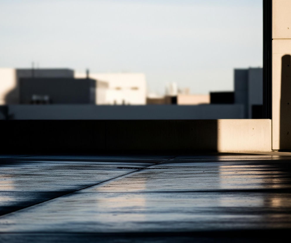 Na pierwszym planie widoczny jest szary, wyłożony prostokątnymi płytami chodnik, częściowo zalany jasnym, złotawym światłem słonecznym tworzącym długie cienie oraz ciemne, nieoświetlone obszary. Po prawej stronie widać fragment beżowej ściany, na którą pada długi, pionowy cień, a dalej w prawo cała przestrzeń jest pogrążona w ciemności. W tle, rozmyte i nieostre, majaczą białe i szare bryły budynków pod jasnym, bezchmurnym niebem.