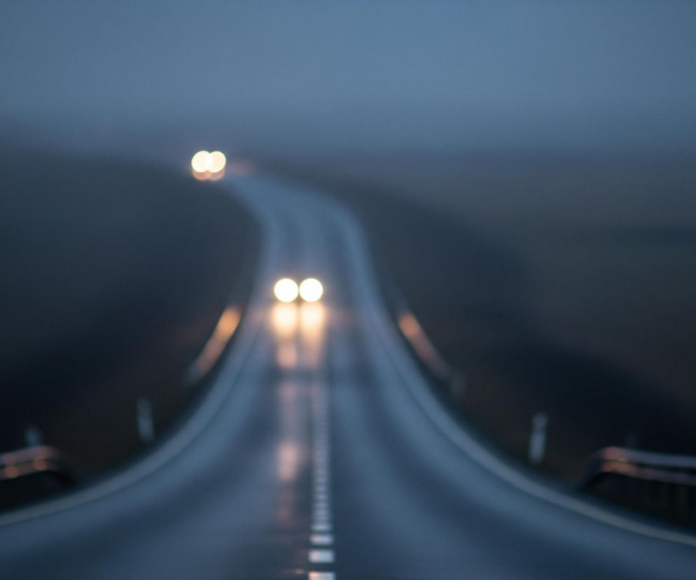 Droga oświetlona światłami samochodu ciągnie się w głąb obrazu, gdzie na zakręcie w oddali widoczne są dwa kolejne, rozmyte światła. Powierzchnia drogi jest mokra i odbija światło, tworząc pionowe smugi i refleksy od przerywanych linii na jezdni. Całe otoczenie, w tym niebo i pobocze, jest spowite w ciemnoszarych i błękitnych tonacjach, co nadaje scenie mglisty, nocny lub przedświtowy charakter.