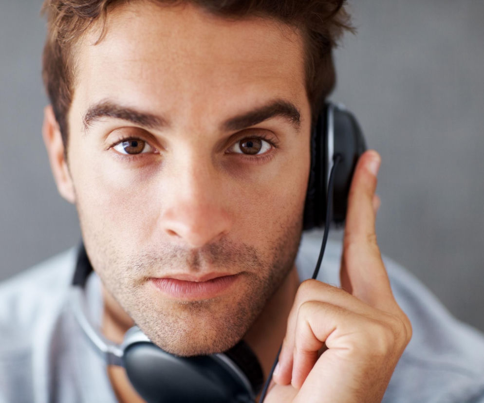 Mężczyzna z brązowymi oczami i ciemnymi włosami, trzymający czarną słuchawkę przy prawym uchu, intensywnie spoglądający wprost. Obraz ilustruje problem zmęczenia i spadku koncentracji przy jednostronnym słuchaniu, o czym przeczytasz na Poradnik Zdrowie.