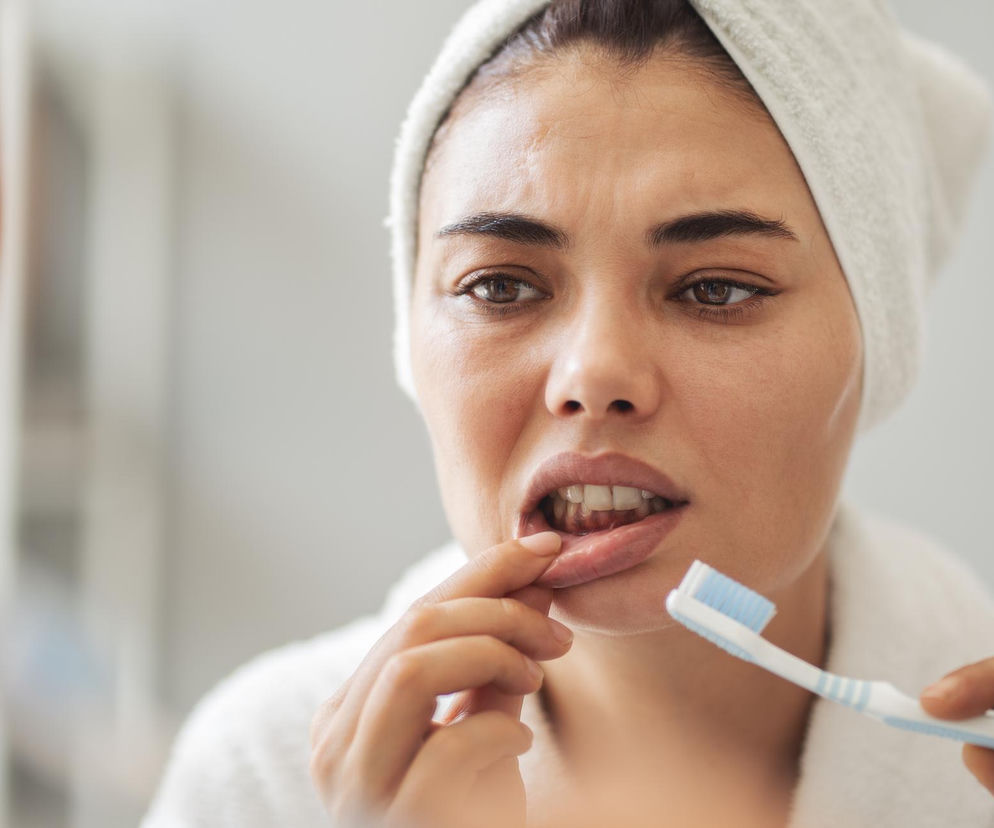 Kobieta w białym ręczniku na głowie i szlafroku, ze szczoteczką do zębów w dłoni, zmartwiona ogląda swoje krwawiące dziąsła przed lustrem. Wskazuje palcem na dolną wargę, co symbolizuje problem z jamą ustną, o którym możesz przeczytać więcej na Poradnik Zdrowie.