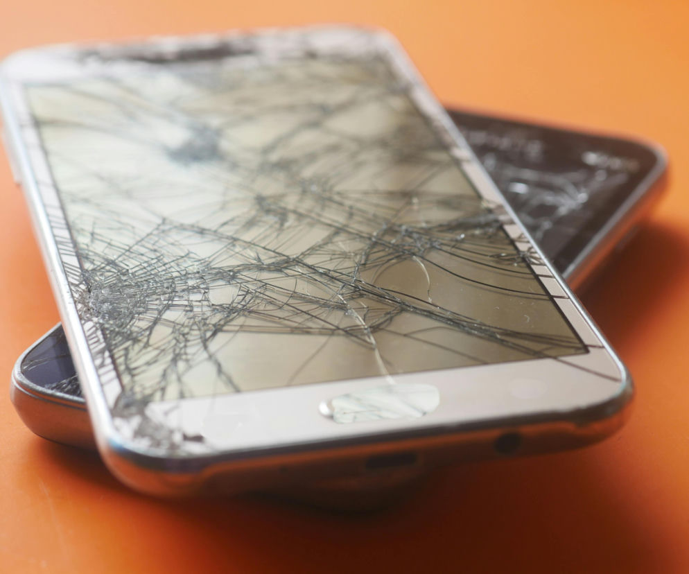 Dwa smartfony z mocno popękanymi ekranami, leżące na pomarańczowej powierzchni, z bliżej widocznym modelem w srebrnej obudowie. Poradnik Zdrowie przestrzega, że pęknięty ekran to idealne siedlisko groźnych bakterii.
