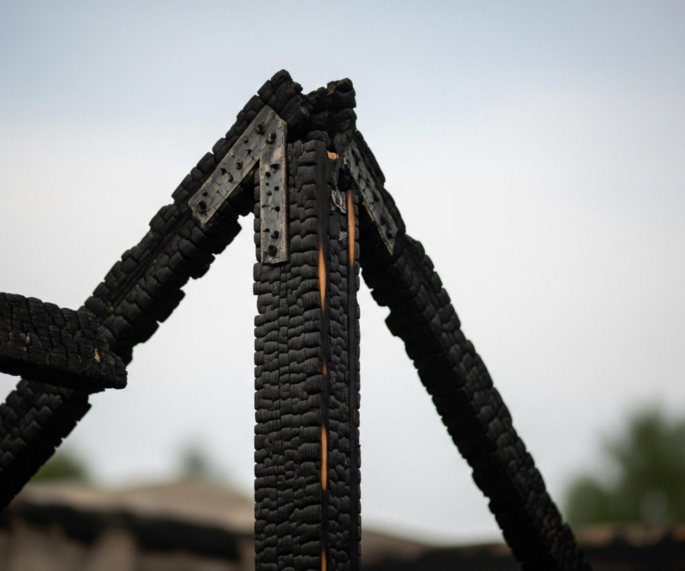 Centralny kadr zajmuje konstrukcja z czterech zwęglonych, czarnych belek drewnianych, połączonych u góry metalowymi elementami z nitami, również częściowo poczerniałymi. Dwie belki rozchodzą się na boki, tworząc rodzaj trójkąta, a jedna z nich jest widoczna w całej długości po lewej stronie, z kolei kolejna biegnie w dół po prawej. Na powierzchni belek widoczne są nierówności i spękania charakterystyczne dla spalonego drewna, z nielicznymi jaśniejszymi, pomarańczowo-brązowymi przebłyskami niezwęglonego drewna. Tło stanowi rozmyte niebo w odcieniach bieli i jasnego błękitu, z niewyraźnymi ciemniejszymi kształtami przypominającymi drzewa i zabudowania w dolnej części.