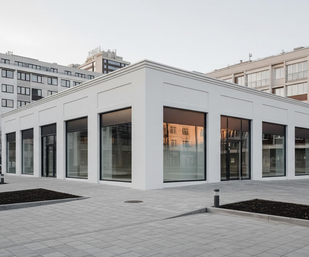 Na pierwszym planie, centralnie, znajduje się niski, prostopadłościenny budynek o białych ścianach i czarnych, dużych oknach bez widocznych zasłon, które odbijają częściowo otoczenie i niebo. Budynek otacza szary chodnik z kostki brukowej, przerywany prostokątnymi rabatami z ciemną ziemią i młodymi drzewami z drewnianymi wspornikami. W tle widoczne są wyższe, wielopiętrowe budynki o różnych fasadach, w tym jeden biały z rzędami czarnych okien po lewej stronie oraz jeden beżowo-brązowy z białymi oknami i balkonami po prawej stronie, wszystko pod jasnym, bezchmurnym niebem.