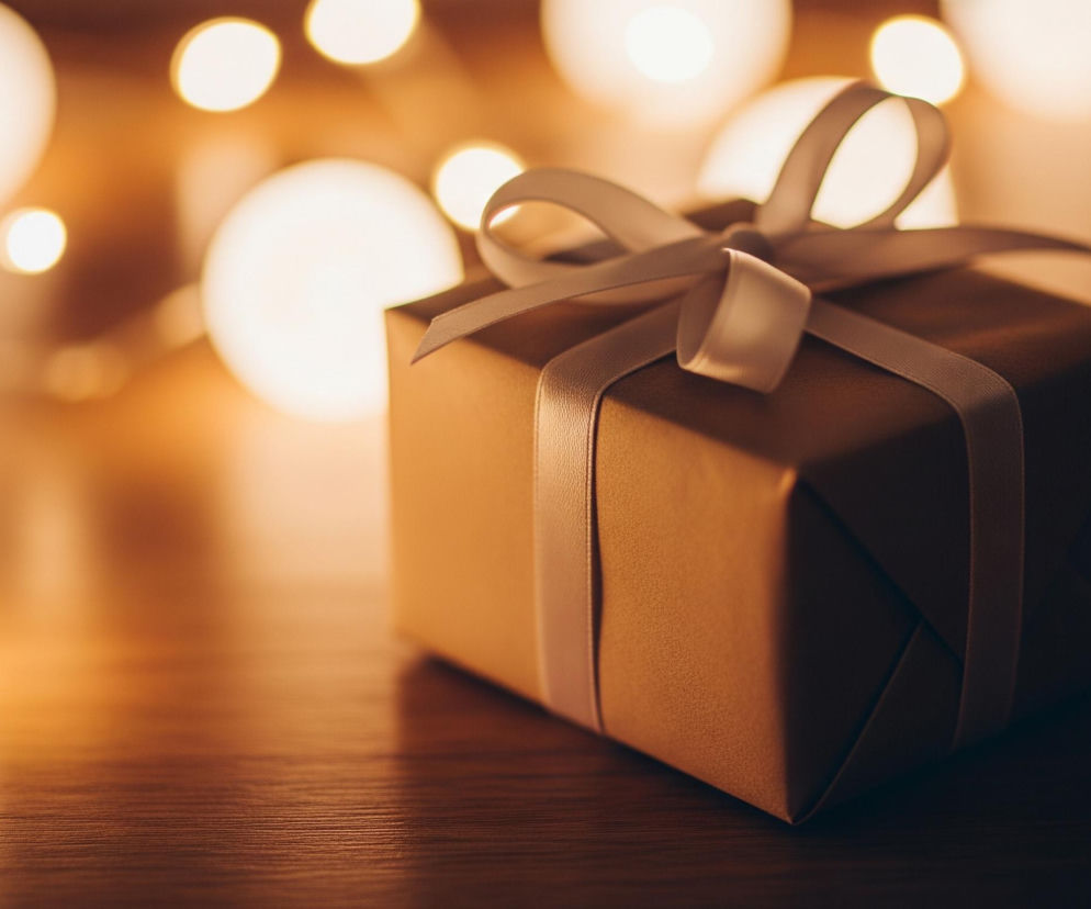 Na pierwszym planie, centralnie, leży brązowe pudełko prezentowe, przewiązane jasną, kremową wstążką, która tworzy ozdobną kokardę na górze. Pudełko spoczywa na ciemnobrązowej, drewnianej powierzchni, prawdopodobnie stole, której faktura jest widoczna. Tło jest rozmyte, ale widać na nim liczne jasne, okrągłe punkty świetlne o ciepłym, żółto-pomarańczowym odcieniu, tworzące efekt bokeh. Całość jest oświetlona ciepłym światłem, co nadaje obrazowi przytulny i świąteczny nastrój.