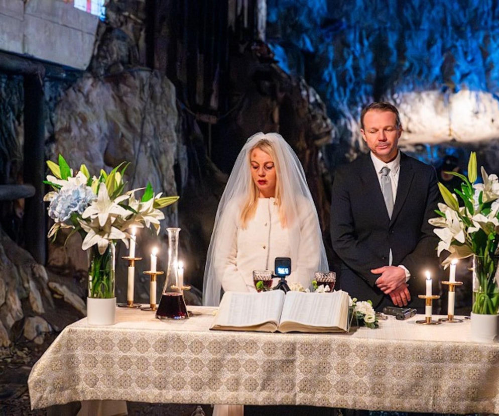Emilka (Anna Ilczuk) w białej sukni ślubnej i welonie z widocznymi siniakami na twarzy, stoi obok Mateusza (Lesław Żurek) w ciemnym garniturze, podczas makabrycznej ceremonii ślubnej w jaskini. Na stole leży otwarta księga, świece i kieliszki z winem, w tle skaliste ściany. Więcej o dramatycznych wydarzeniach w serialu Pierwsza miłość przeczytasz na portalu Super Seriale.