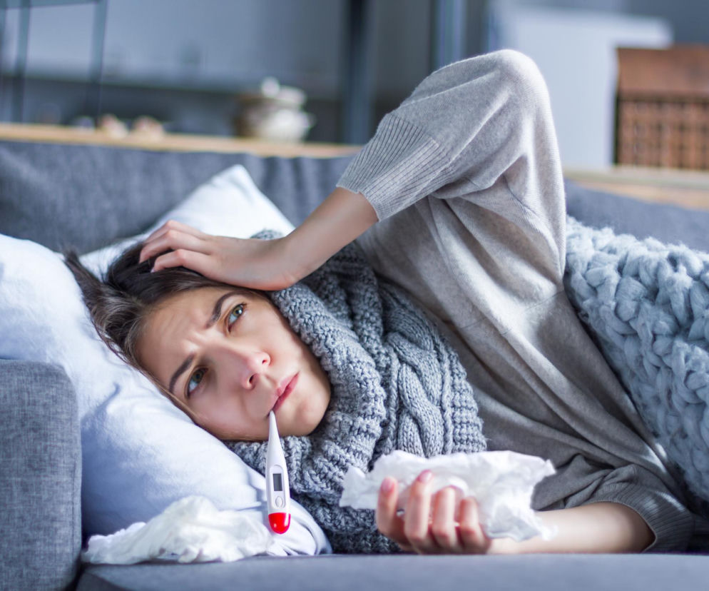 Zmęczona kobieta z termometrem w ustach leży na kanapie, okryta szarym kocem, odczuwając dreszcze i zimno. Obok niej leżą zużyte chusteczki. Na Poradnik Zdrowie można przeczytać o tym, co mózg robi, gdy mamy dreszcze przy gorączce.