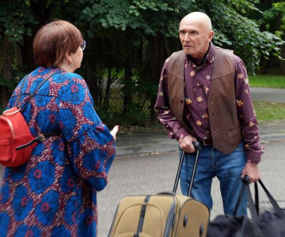 Irenka (Krystyna Tkacz) w niebieskiej sukience z czerwonymi wzorami i czerwoną torebką rozmawia z Tolkiem (Marek Siudym), który w koszuli, kamizelce i jeansach, z walizkami, opuszcza dom. Zdjęcie przedstawia dramatyczne rozstanie bohaterów serialu Barwy szczęścia, o którym przeczytasz na portalu Super Seriale.