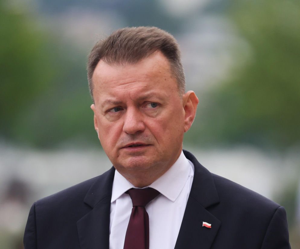 Mariusz Błaszczak w granatowym garniturze z białą koszulą i bordowym krawatem, z przypinką polskiej flagi na klapie marynarki. Polityk patrzy w bok, a jego wyraz twarzy jest poważny. Decyzję Błaszczaka o zwrocie orderu za zasługi, przeczytasz na naszym portalu.