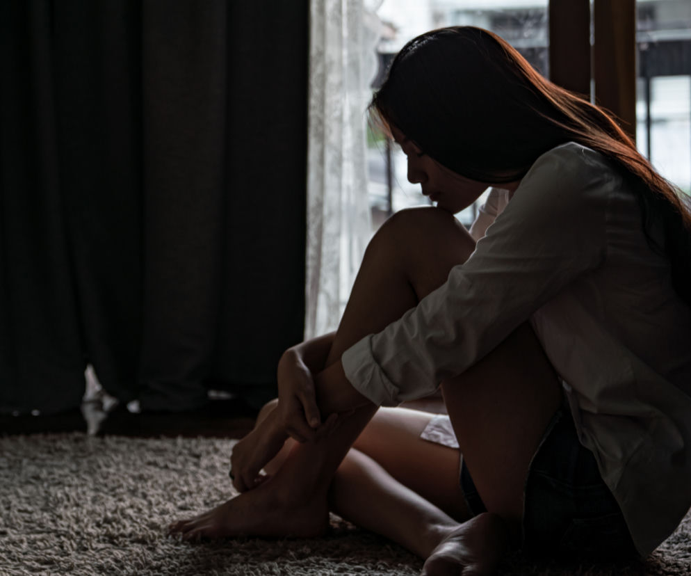 Młoda kobieta, nastolatka, siedzi skulona na podłodze, symbolizując trudną sytuację oddziału psychiatrycznego dla młodzieży w Konstancinie. Jej smutna, zamyślona postawa odzwierciedla emocje młodych pacjentów po próbach samobójczych. Więcej o kryzysie psychiatrii dziecięcej przeczytasz na Poradnik Zdrowie.